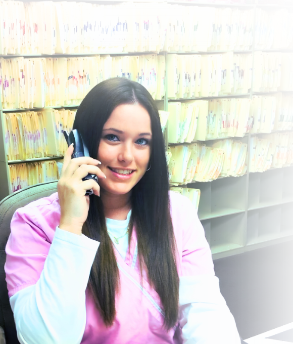 lady sitting at a desk with files behind her wearing a nurse uniform talking on the phone with a smile on her face. she has long hair and light skin